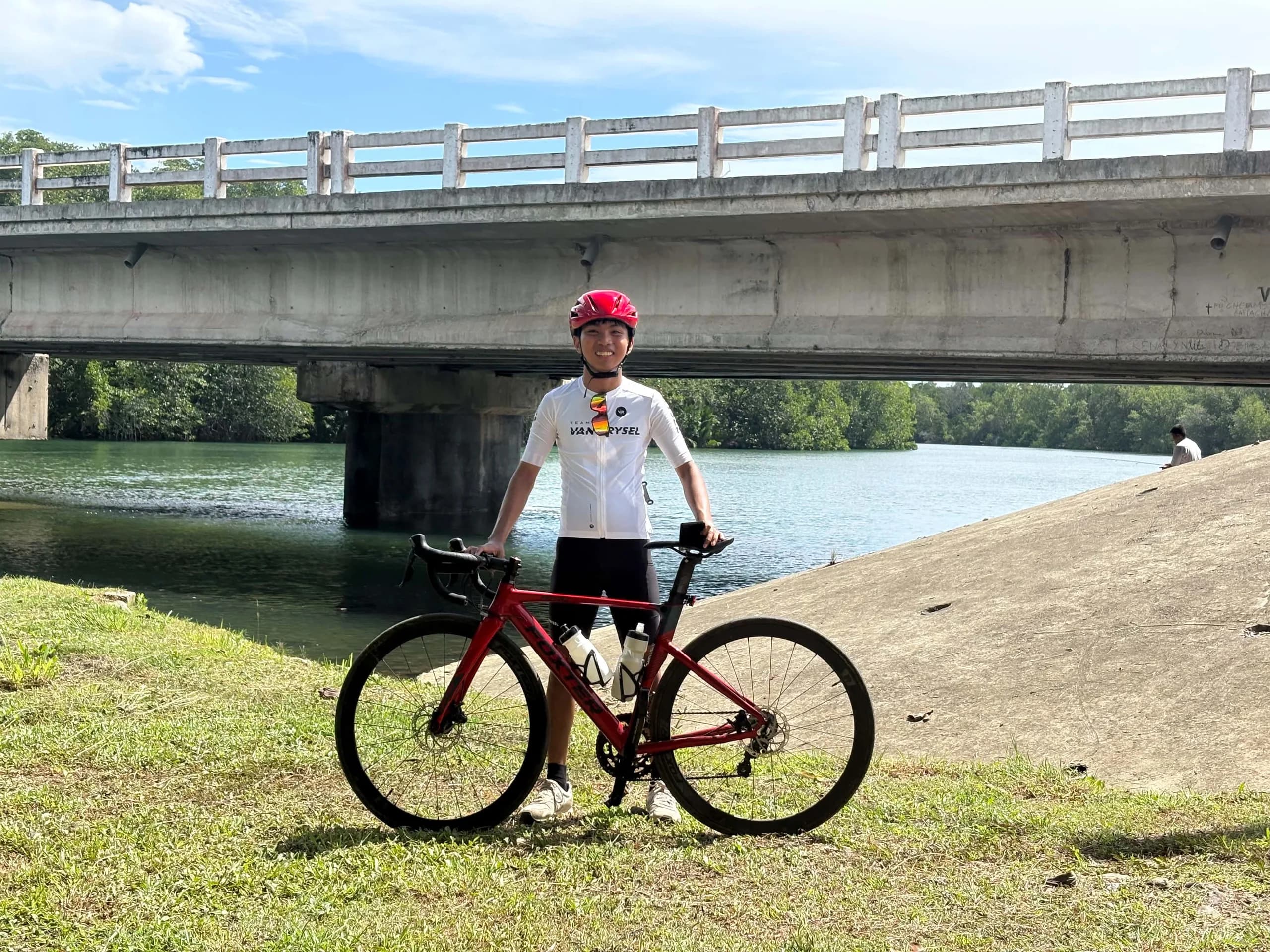 Elsid standing with a red road bike on a grassy riverbank below a concrete bridge