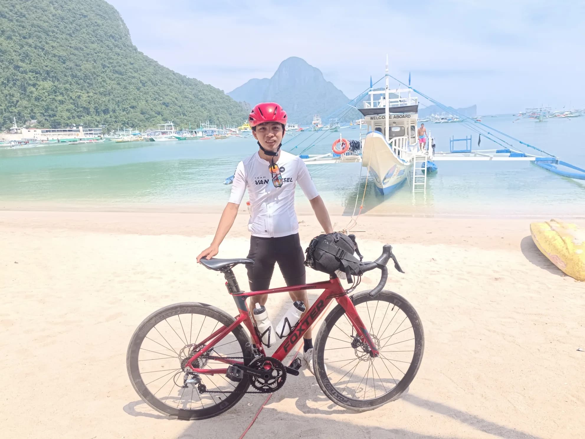 Elsid with a red road bike on the beach in El Nido, with bangka boats and limestone cliffs behind