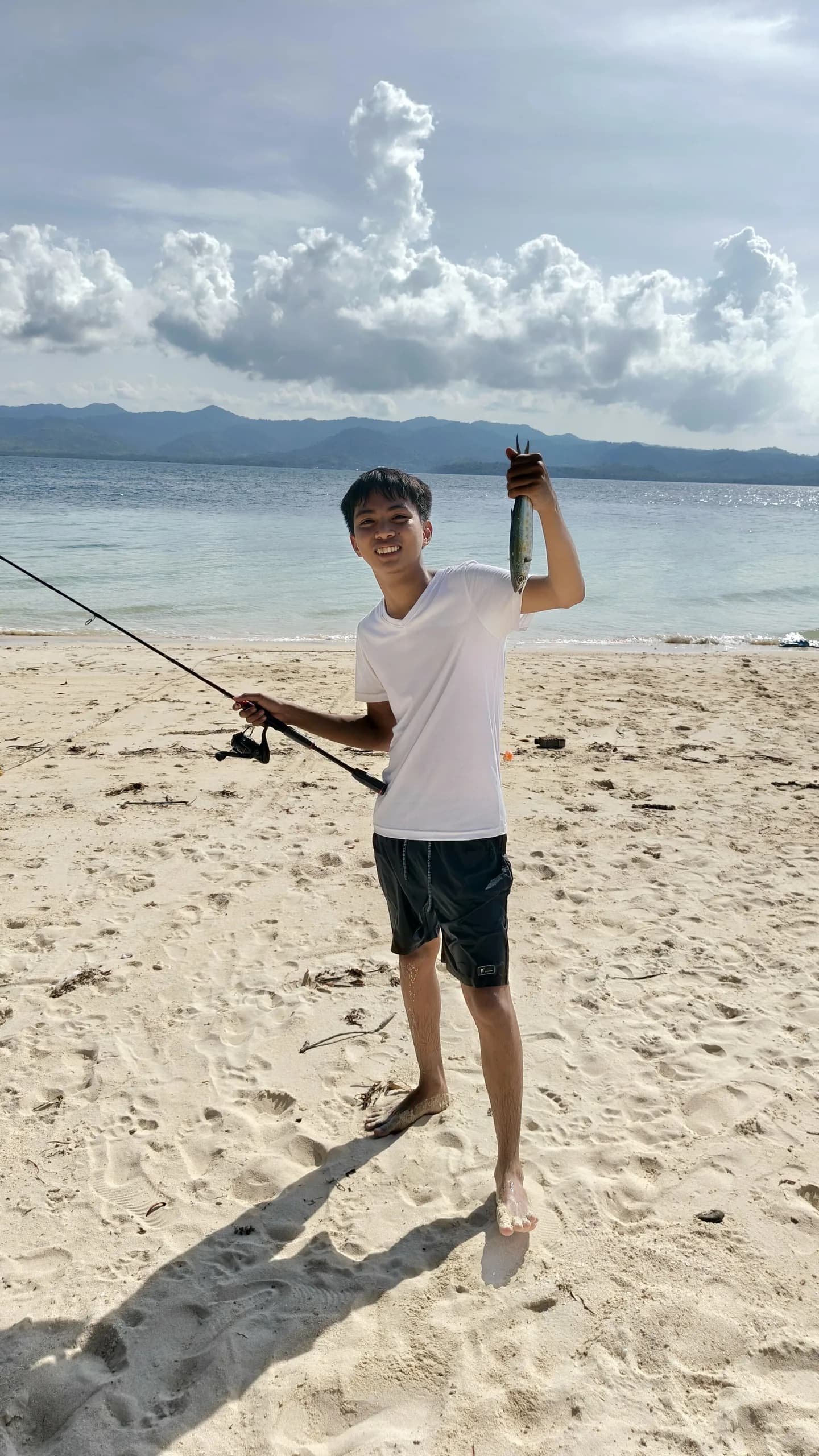 Elsid on a sandy beach in Taytay holding up a small fish, with the sea and mountains behind
