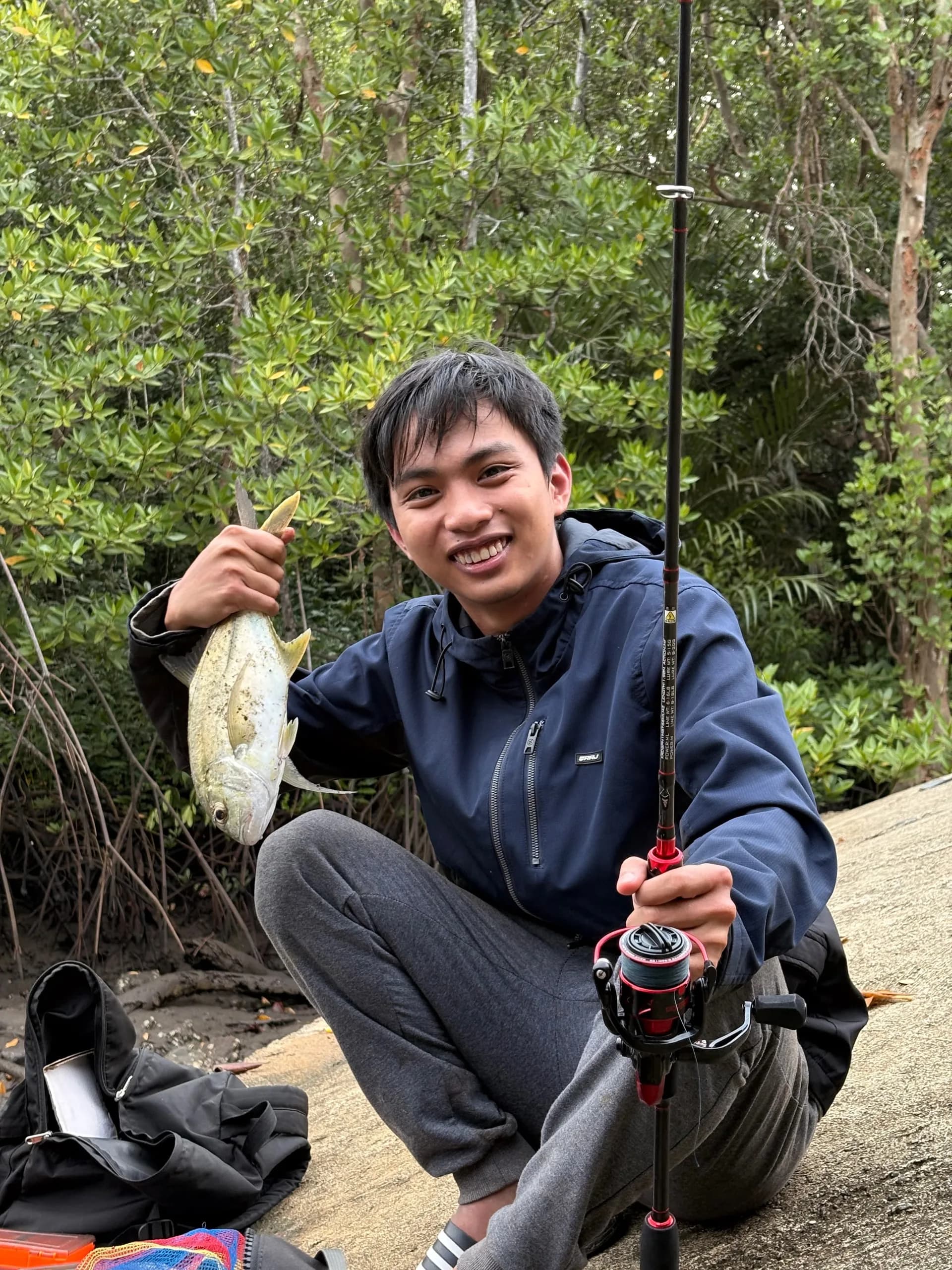 Elsid crouching by the mangroves, holding up a silver fish in one hand and a fishing rod in the other