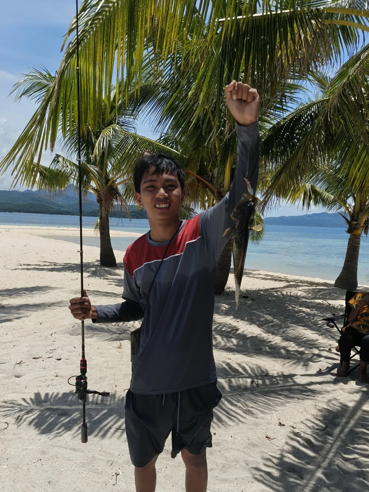 Elsid on a palm-lined white-sand beach in Taytay, raising a fish and holding a fishing rod